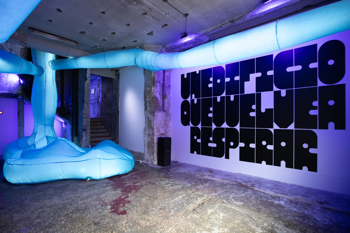 Sala con techo de hormigón picado, tubos celestes y una pared blanca con una frase pintada.